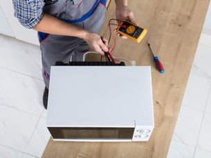 Technician tests a microwave with a multimeter on a wooden counter, with a screwdriver nearby.