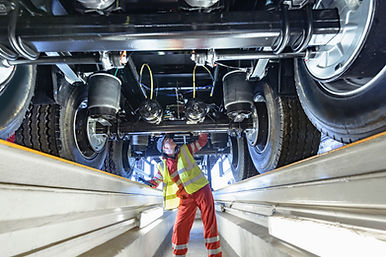 A mechanic in a high-visibility vest and red overalls inspecting the underside of a large vehicle