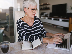 A woman with short gray hair and glasses types on a laptop at a table. An open notebook and a glass of water are nearby in a cozy room.