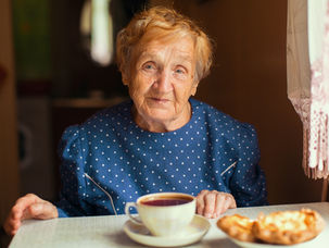 Elderly woman in blue polka-dot dress sits at a table with a cup of tea and pastries, softly lit, expressing warmth and contentment.