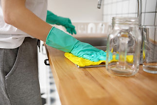 Person wearing green rubber gloves wipes a wooden kitchen counter with a yellow cloth