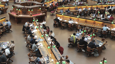 Four students walk down a spiral staircase with orange railings, holding backpacks and books, in a building with gray steps and a modern design.
