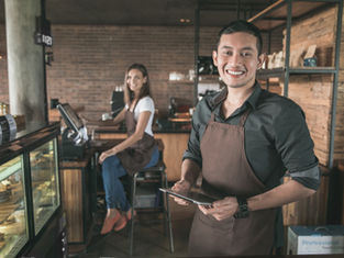 Two smiling baristas in brown aprons inside a café. One holds a tablet, the other sits at a counter. Brick wall and coffee station visible.