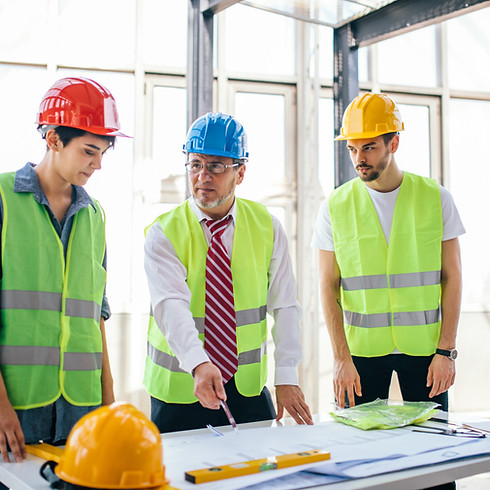 Three construction workers in hard hats and safety vests review blueprints on a table