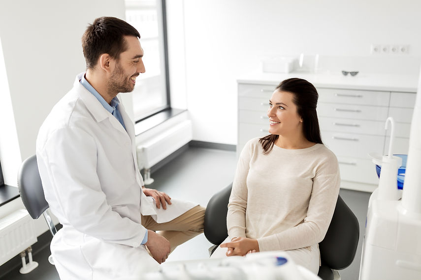 A dentist in a white coat smiles at a seated woman in a dental office