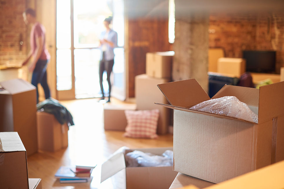 An open cardboard moving box with bubble wrap in focus, with a couple standing out of focus near the sunlit balcony doorway