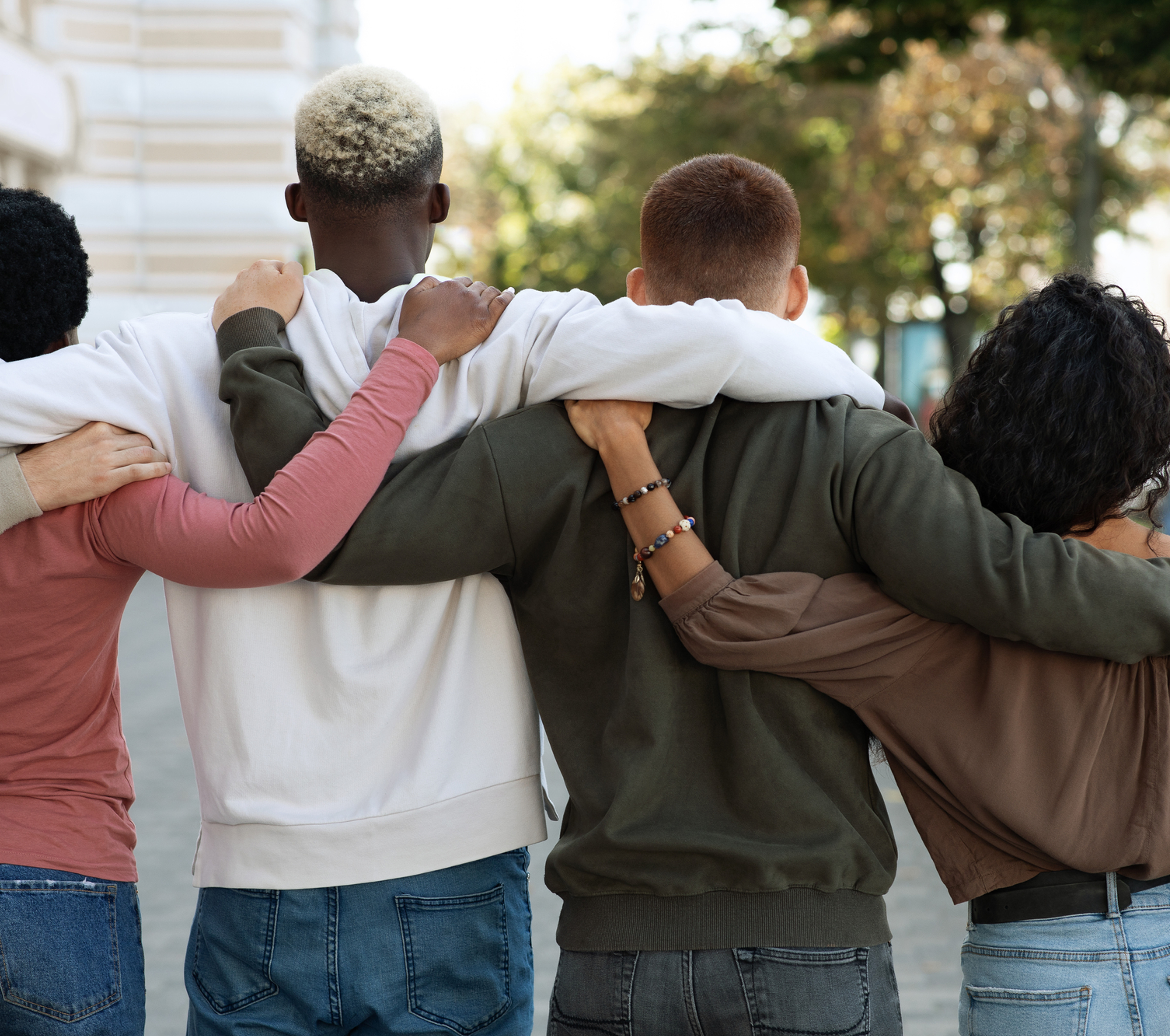 Four friends with arms around each other walk down a sunlit street, seen from behind, showing unity and support.