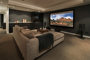 A cozy home theater with a large beige sectional sofa facing a big screen showing a desert landscape