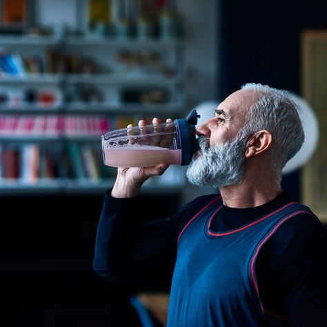 Man in blue sports vest drinks pink smoothie in kitchen with bookshelves, looking content. Dark, cozy background adds warmth.