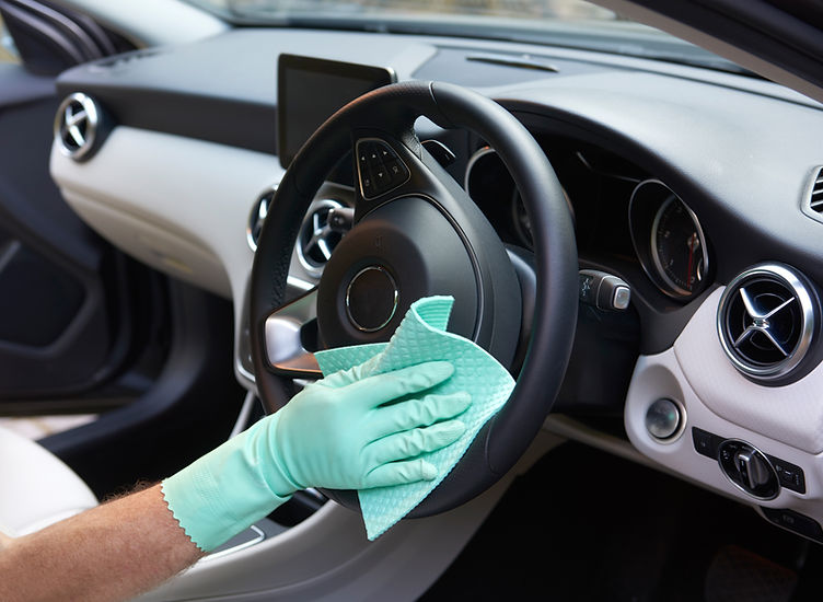 A person wearing teal gloves cleans a car's steering wheel with a cloth inside a modern vehicle