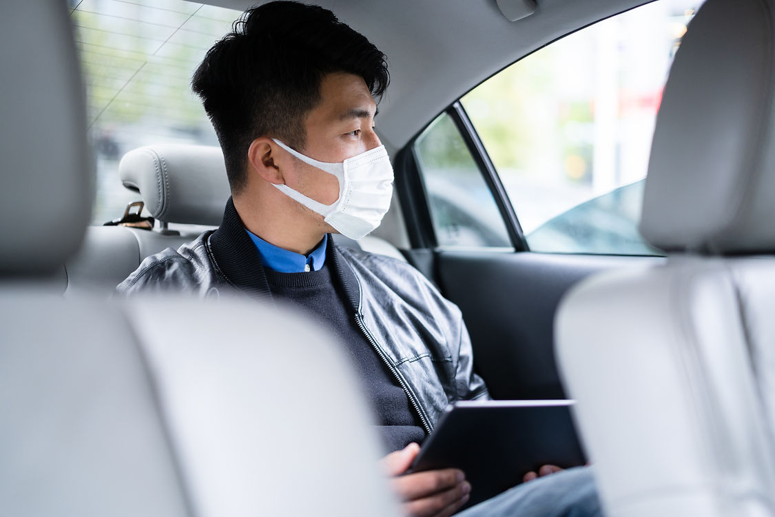 A man wearing a face mask sits in the back seat of a car, looking out the window