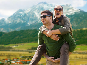 Smiling couple in sunglasses, woman riding piggyback on man, with snowy mountains and green valley behind them.