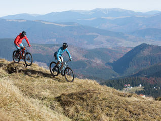 Two mountain bikers in red and blue ride along a grassy ridge above a vast mountain valley.