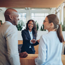 Smiling woman hands a card to a couple in an office lobby. Another woman works at a computer, talking on the phone. Bright, modern setting.