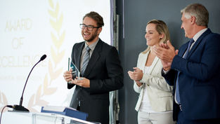 Man in a suit holds an award, smiling. Two colleagues clap beside him. "Award of Excellence" text in the background. Professional setting.