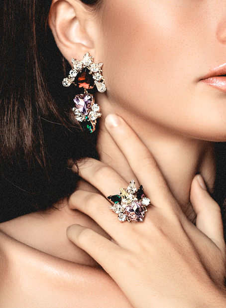 Woman wearing ornate earrings and ring with colorful gems, touching her neck. Close-up shows rich colors and elegance.