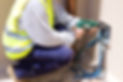 A worker in a high-visibility vest and blue trousers using a green power drill on a wall