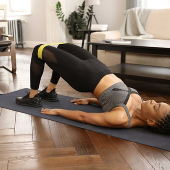 Woman in activewear doing a glute bridge on a mat in a living room. A laptop nearby displays an exercise guide. Bright, cozy setting.