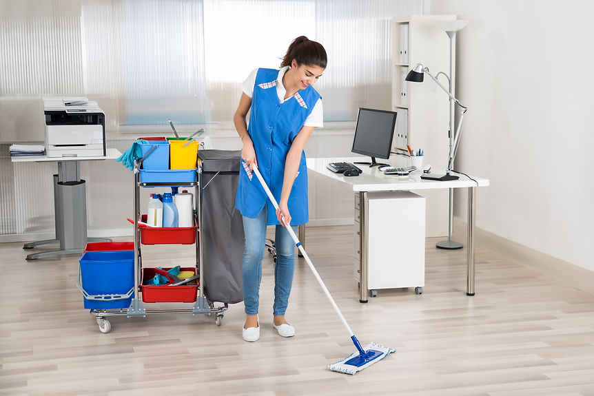 A woman in blue uniform mops a bright, tidy office with a cleaning cart nearby