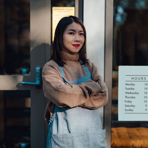 Woman in a tan sweater and gray apron stands confidently with arms crossed outside a shop. Hours are posted on the door. Mood is determined. Image implies authentic confidence.