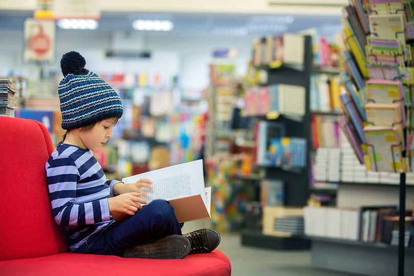 Child Reading Book. Wearing blue stripey top and blue bobble hat. They are in a bookshop.