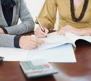 Two people collaborate at a table covered with papers, one writing with a pen.