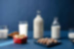 Bottles and glasses of milk on a blue background, surrounded by almonds, hazelnuts, and cashews in red and blue dishes.
