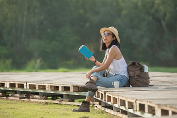 Woman in hat and sunglasses sits on wooden platform outdoors, holding a blue book, surrounded by nature. Backpack and mug nearby. Smiling.