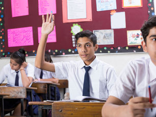 schoolboy in white shirt and tie raises hand in classroom while classmates write at desks; colorful bulletin board behind.