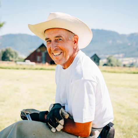 Man in white shirt and cowboy hat sitting on grass, smiling. Gloves in hand. Sunny farm setting with barn and trees in background.