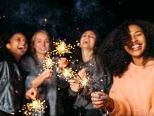 Four people joyfully hold sparklers at night, their faces lit with smiles. Sparks illuminate the dark background, creating a festive atmosphere.