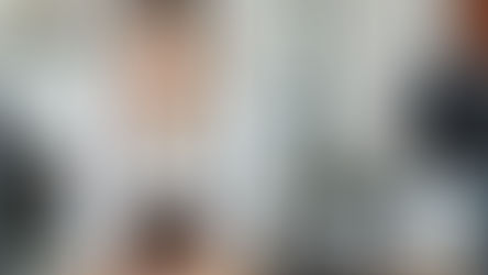 Barista in a white shirt and brown apron smiles, leaning on a counter. Jars and a register are present. The setting is a bright café.