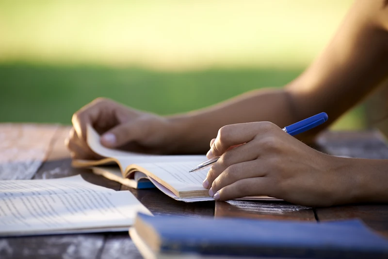A pair of hands on top of a book, the offscreen person appears to be reading and taking notes in an outdoor space.