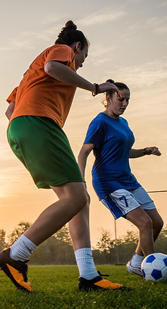 Two women playing soccer on a grassy field at sunset.