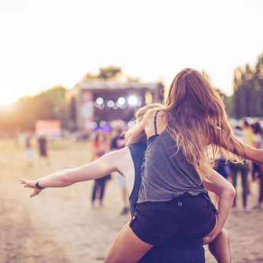 Young woman rides on someone’s shoulders with arms outstretched at a sunset music festival, crowd and stage blurred behind.