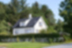 A white wooden house surrounded by overgrown green shrubs and a white fence.