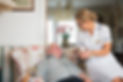 A caregiver in a white uniform and glasses smiles warmly at an elderly man seated in a floral armchair