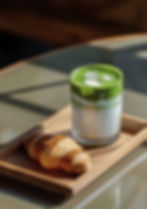 Iced matcha latte and croissant on a wooden tray, bathed in warm sunlight. Green and white layers with a cozy, inviting mood.