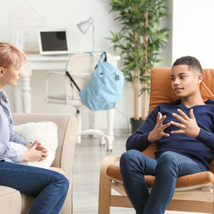 Life coach talks with a teen in a bright office, with a laptop, lamp, plant, and backpack in the background.
