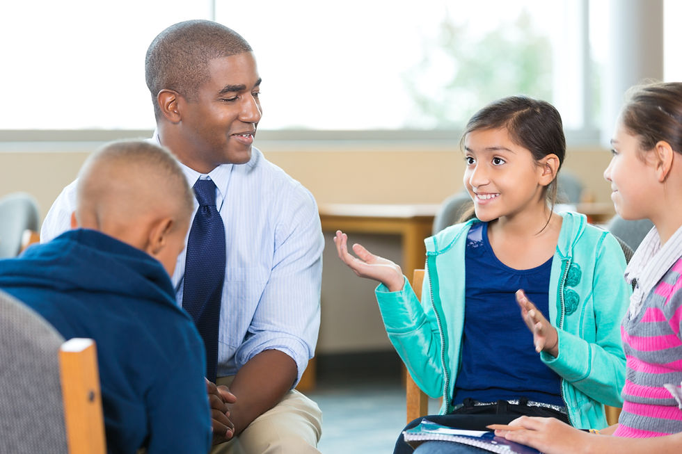 A teacher and three kids sit together, smiling and conversing in a classroom. The girl in teal gestures animatedly, creating a cheerful mood.