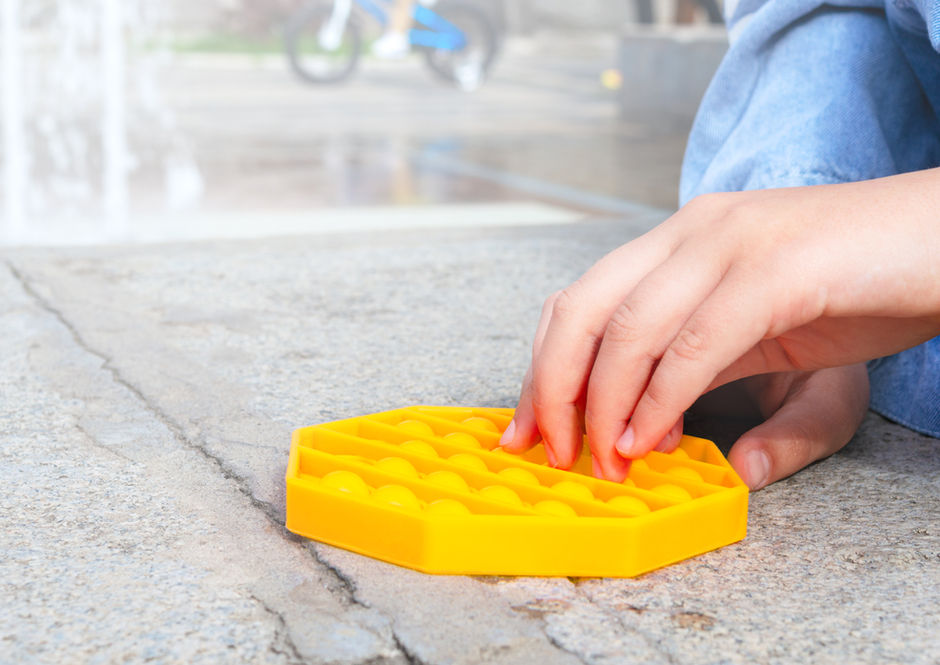 Child's hand presses yellow hexagonal pop-it toy on concrete pavement.