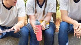 Three students in white shirts and jeans sit outdoors on a wall, one holding a pink water bottle and another a tablet.