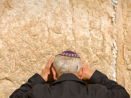 Man in dark coat praying at a stone wall with notes in cracks, wearing a purple kippah. The scene conveys a peaceful mood.