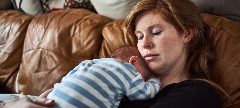 A close up of a woman sitting on a leather sofa, she is holding a baby on her chest, her head is leaned it the baby's head and her eyes are closed.