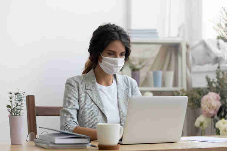 This is a picture of a woman sitting at a table in front of an open laptop. She's in a small, white room, and she's wearing a light colored shirt, a gray blazer, and a white mask across her face. Beside her is a white coffee mug, some notebooks, a pair of glasses, and a plant. In the background is a bookshelt, a window, and a large plant. 