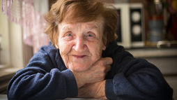 Smiling elderly woman in a blue sweater sitting indoors with her arms crossed on a table, representing confidence, comfort, and positive senior health.