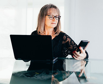 Older woman looking at her phone with a smile while seated at her computer, representing real-time text-based home organizing support for women