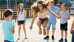 Children enjoying gross motor activities as they play jump rope together on a sunny playground, with one girl jumping while others watch nearby.