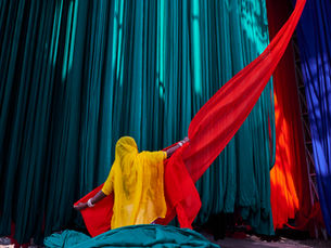 A person in a bright yellow garment holds flowing red fabric against a backdrop of hanging teal and blue textiles, creating a vibrant scene.