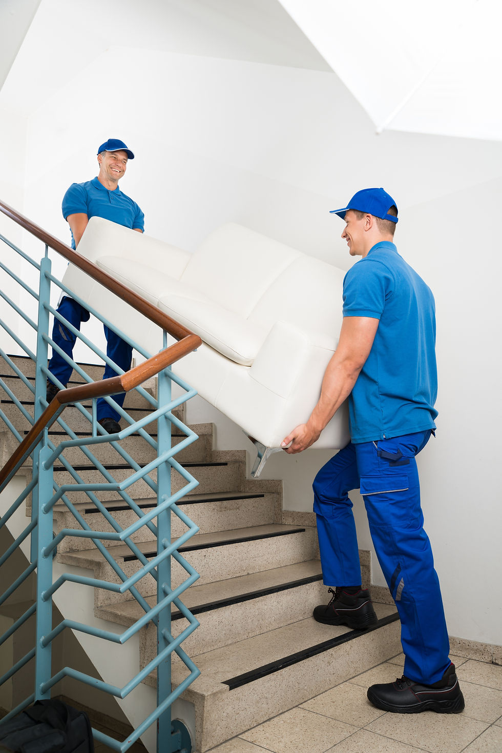 two people in blue uniforms carrying a white sofa up a staircase